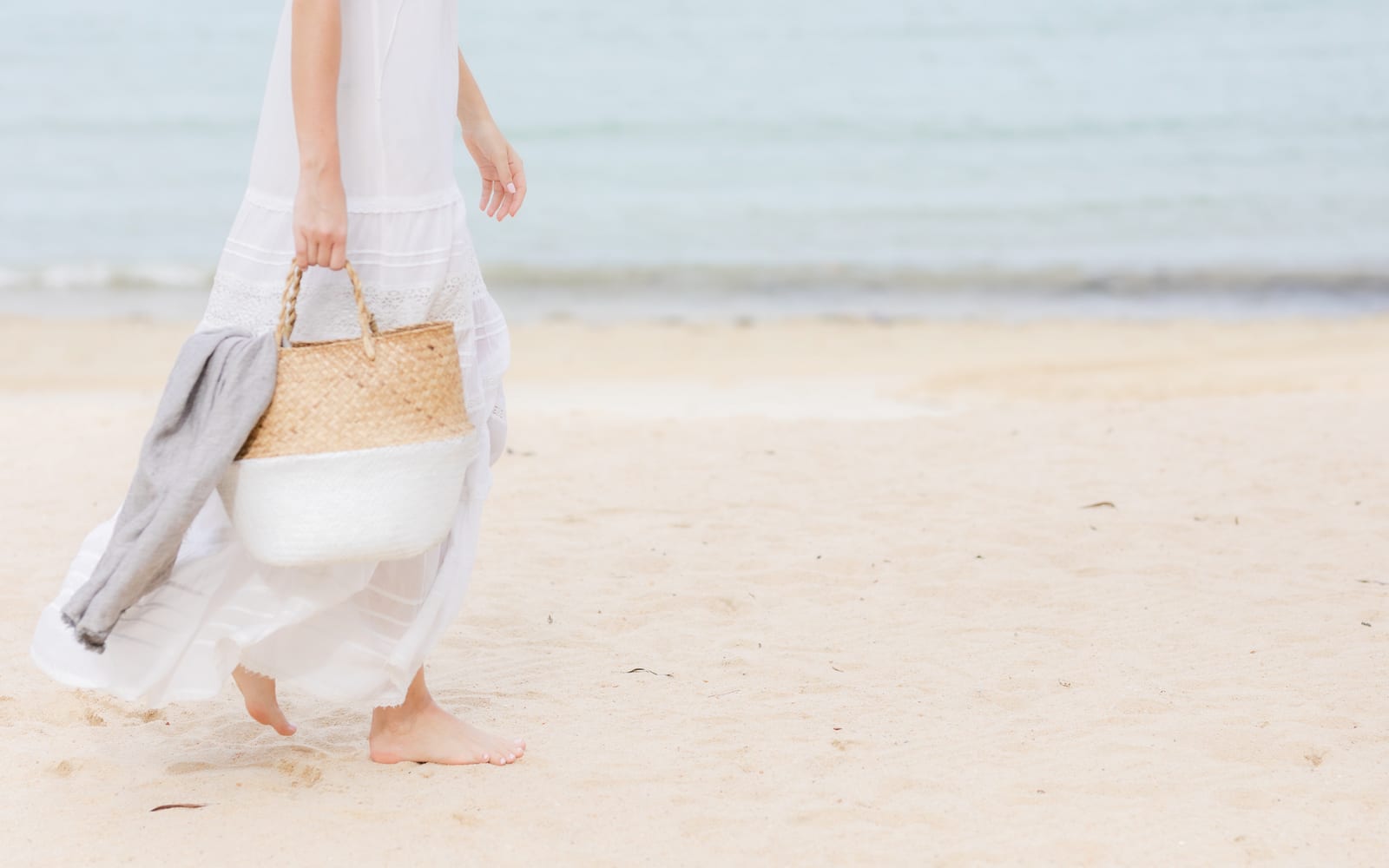 Frau mit Strandtasche geht barfuß am Meer entlang, Symbolbild für Verstopfung im Urlaub
